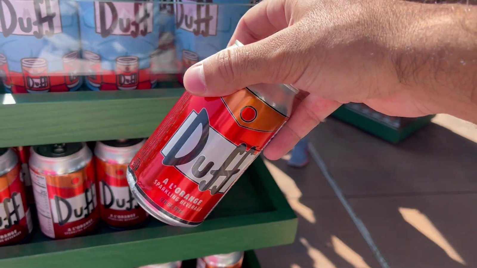 Latinha vermelha de Duff na mão, em frente à prateleira de latas de Duff na loja de Springfield