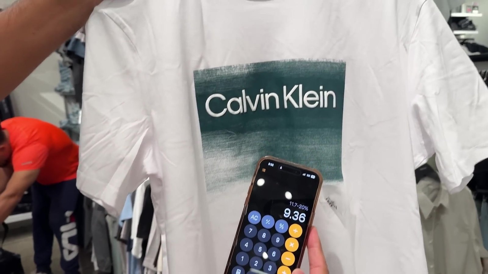 Camiseta Calvin Klein no outlet com a calculadora do celular mostrando US$ 9,36 depois dos descontos