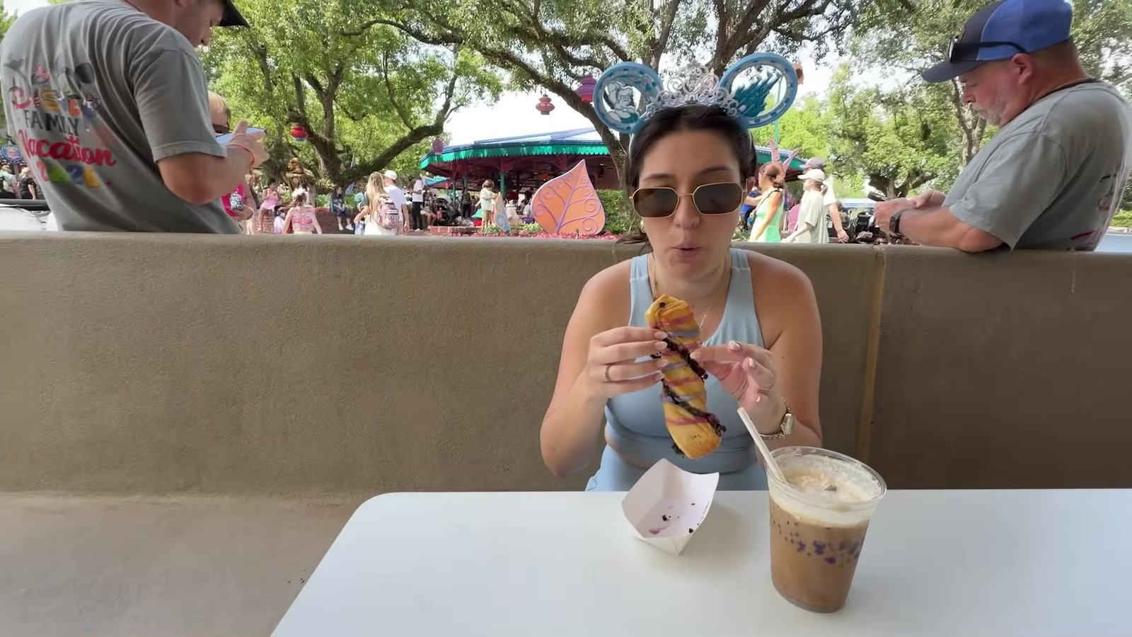 Gabi sentada numa mesa do Magic Kingdom mordendo o croissant de chocolate com um café gelado ao lado