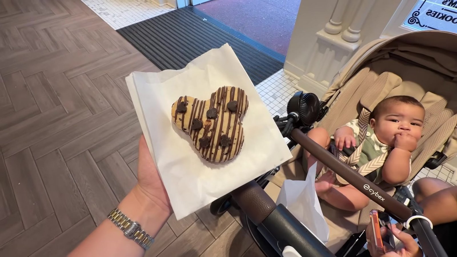 Doce em formato de Mickey com cobertura de chocolate na mão, ao lado do carrinho de bebê