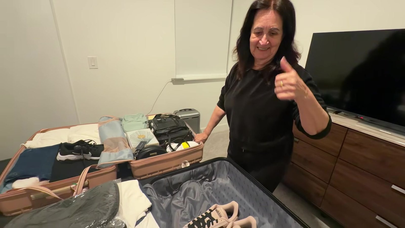 Mãe da Gabi fazendo joinha ao lado da mala aberta na cama