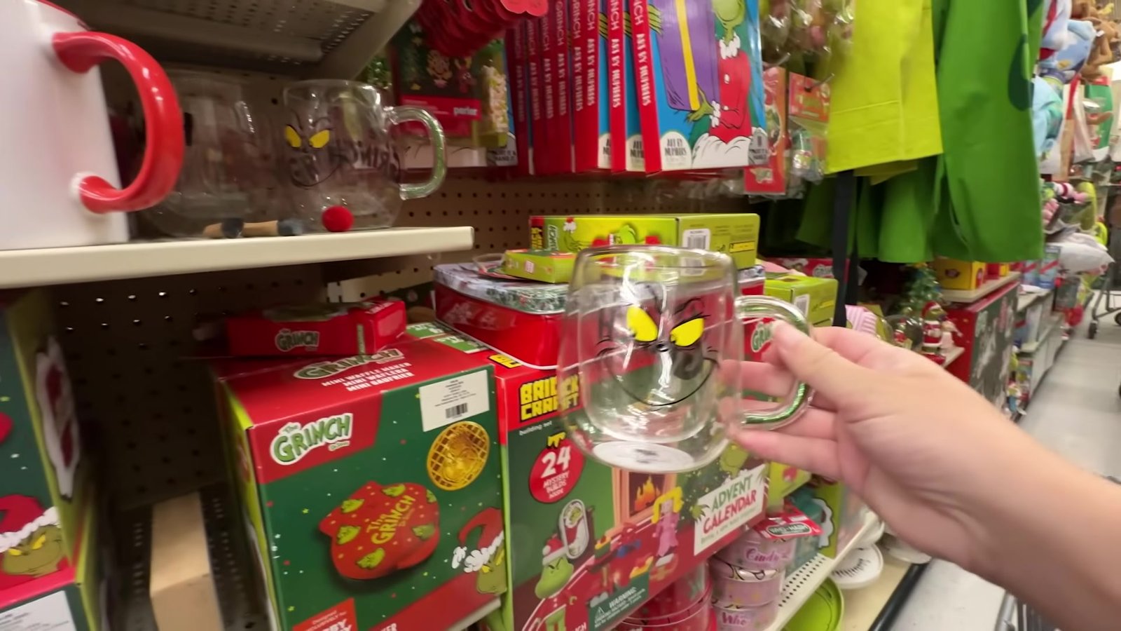 Caneca de vidro do Grinch na mão, com caixas de chocolate quente do Grinch na prateleira atrás