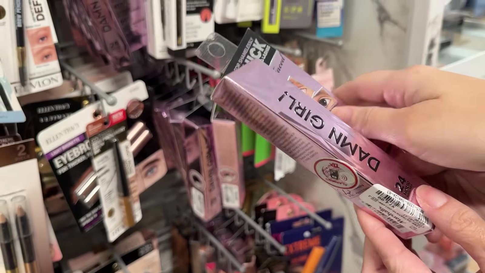 Mão segurando a caixa roxa do rímel Damn Girl da Too Faced na gôndola de maquiagem da Marshalls