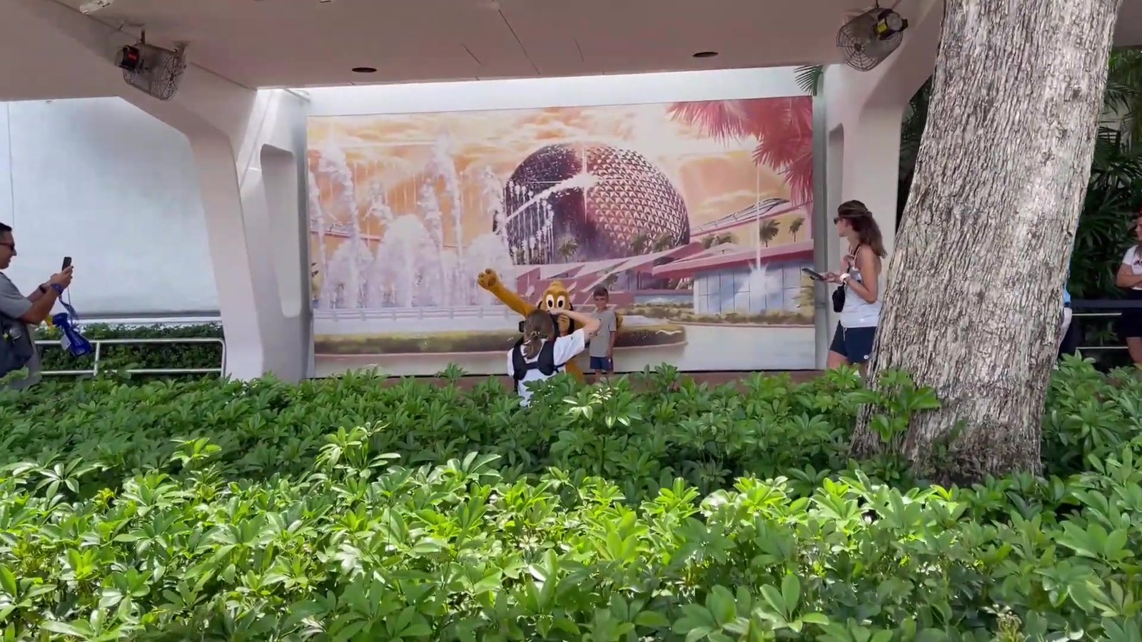 Pluto posando com visitantes em frente ao mural do Epcot, ponto de foto do PhotoPass