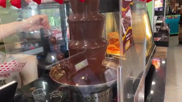 Espetinho de morango sendo banhado na fonte de chocolate da Cantina Brazil, com as pizzas ao fundo