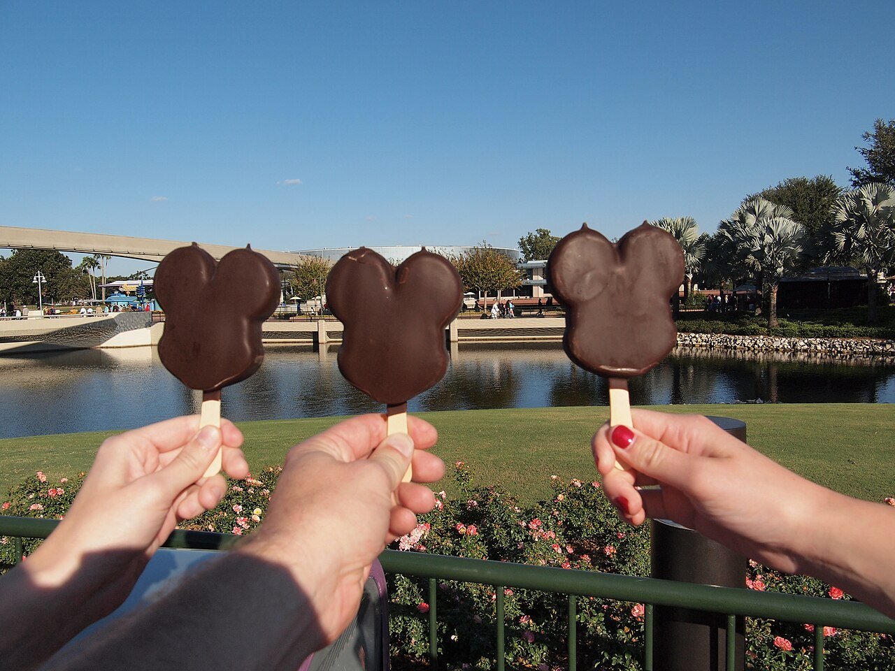 Três picolés do Mickey de chocolate segurados em frente ao lago do Magic Kingdom