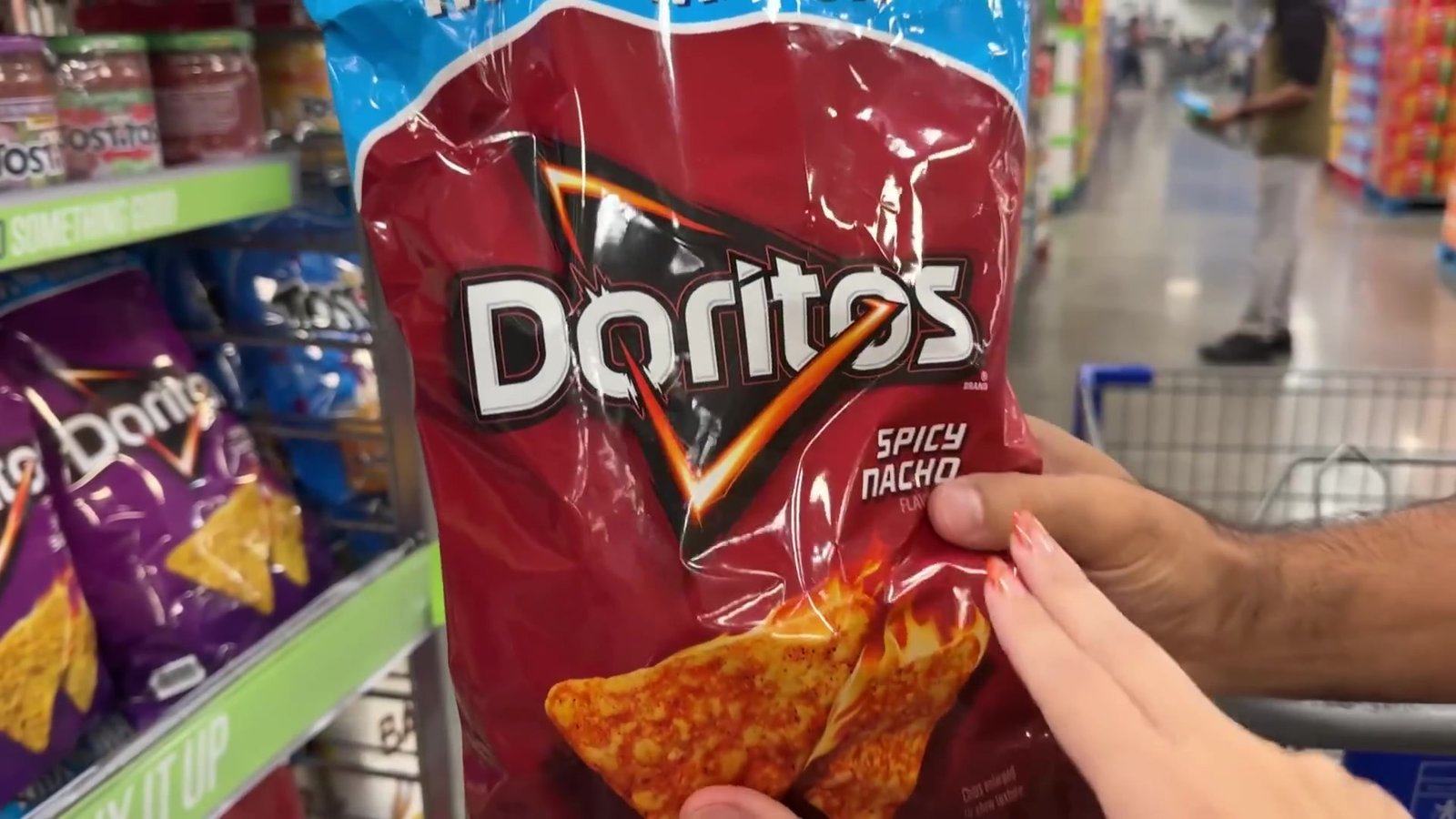 Gabi segurando o saco gigante de Doritos Spicy Nacho no corredor de salgadinhos do Sam's Club