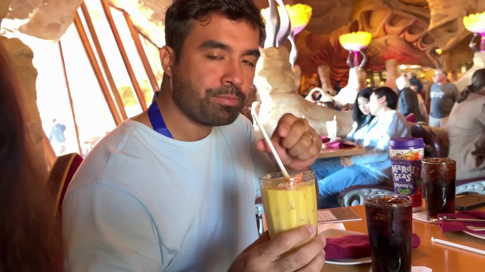 Gustavo tomando um drink amarelo no salão do Mythos, com as paredes de rocha esculpida ao fundo